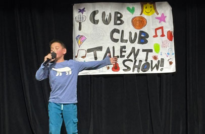 Boy speaking into a microphone on stage in front of a sign that says Cub Club Talent Show